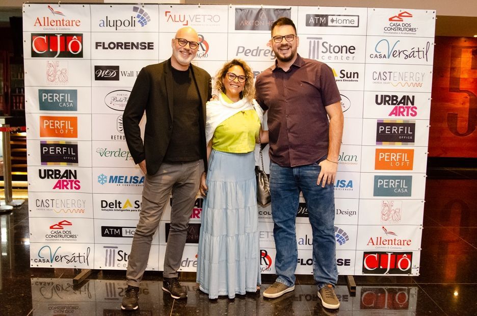 Três pessoas estão posando para uma foto em frente a uma parede com logotipos.