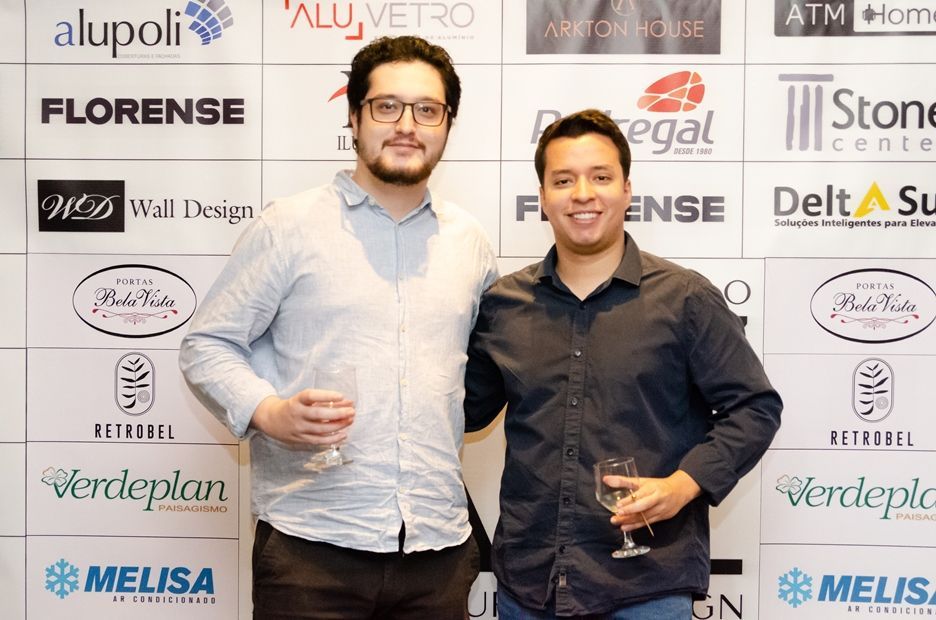 Dois homens estão posando para uma foto em frente a uma parede com logotipos.