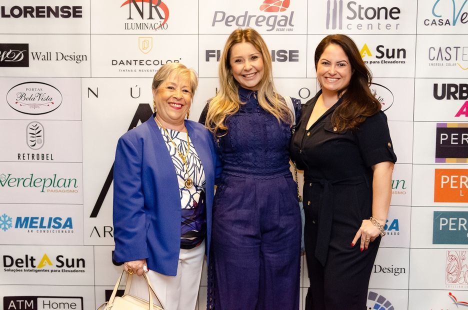 Três mulheres posam para uma foto em frente a uma parede com logotipos de empresas como a pedregat