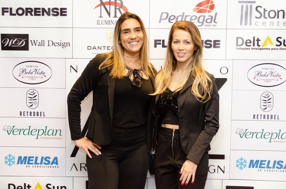 Duas mulheres estão posando para uma foto em frente a uma parede com logotipos.