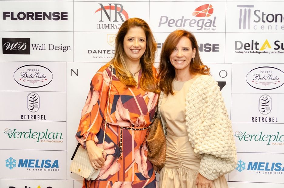 Duas mulheres estão posando para uma foto em frente a uma parede com logotipos.