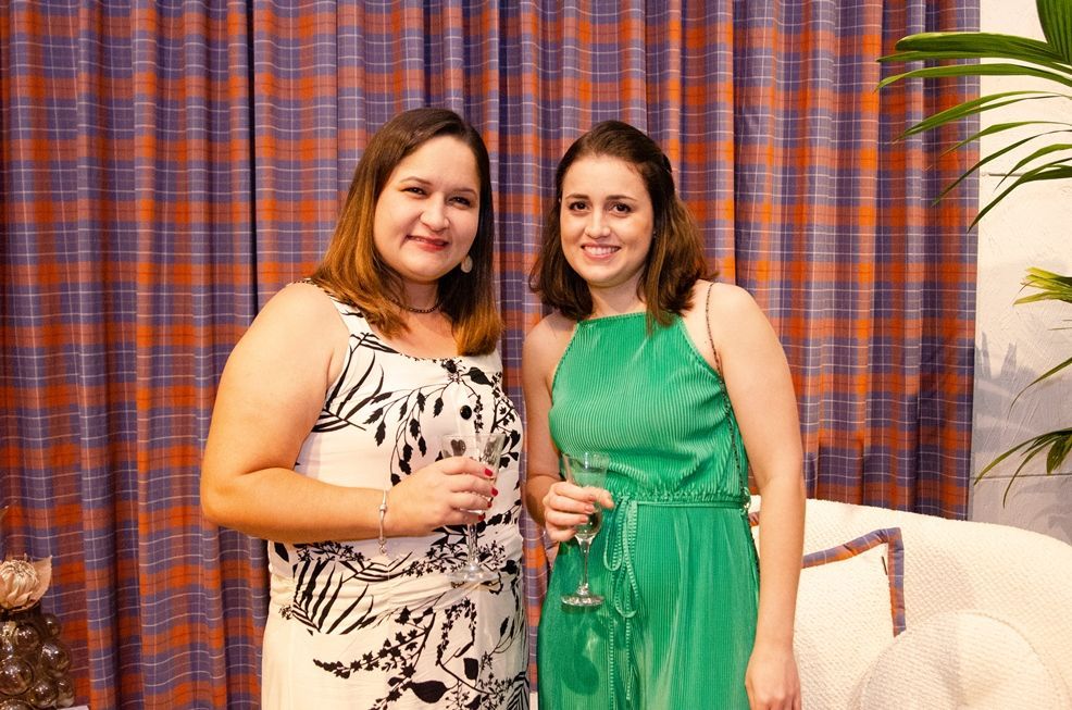 Duas mulheres estão de pé, uma ao lado da outra, em frente a uma cortina, segurando taças de vinho.