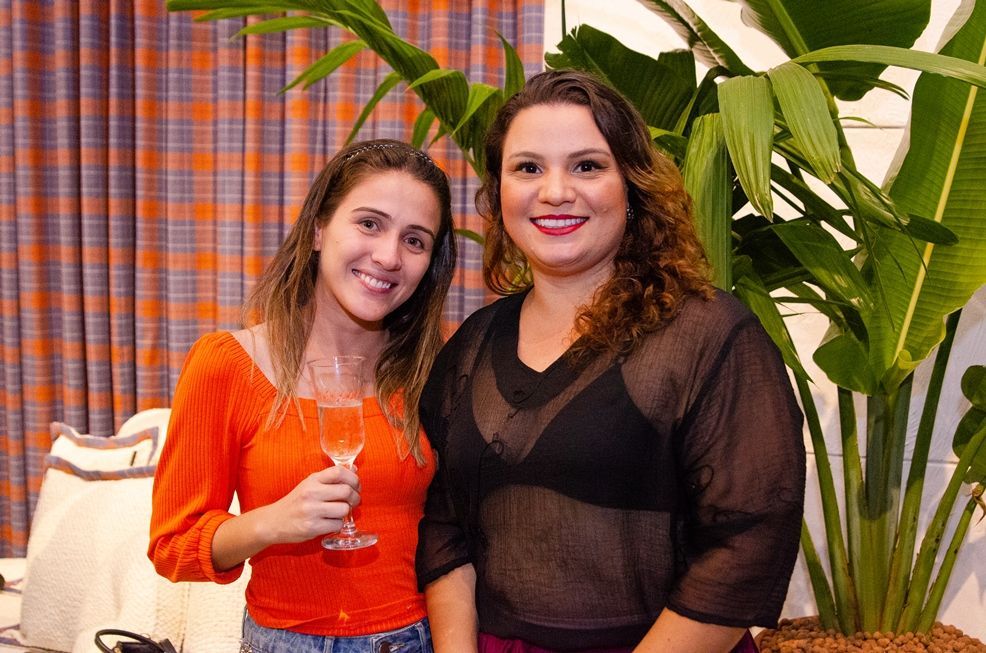 Duas mulheres estão de pé, uma ao lado da outra, segurando taças de vinho.