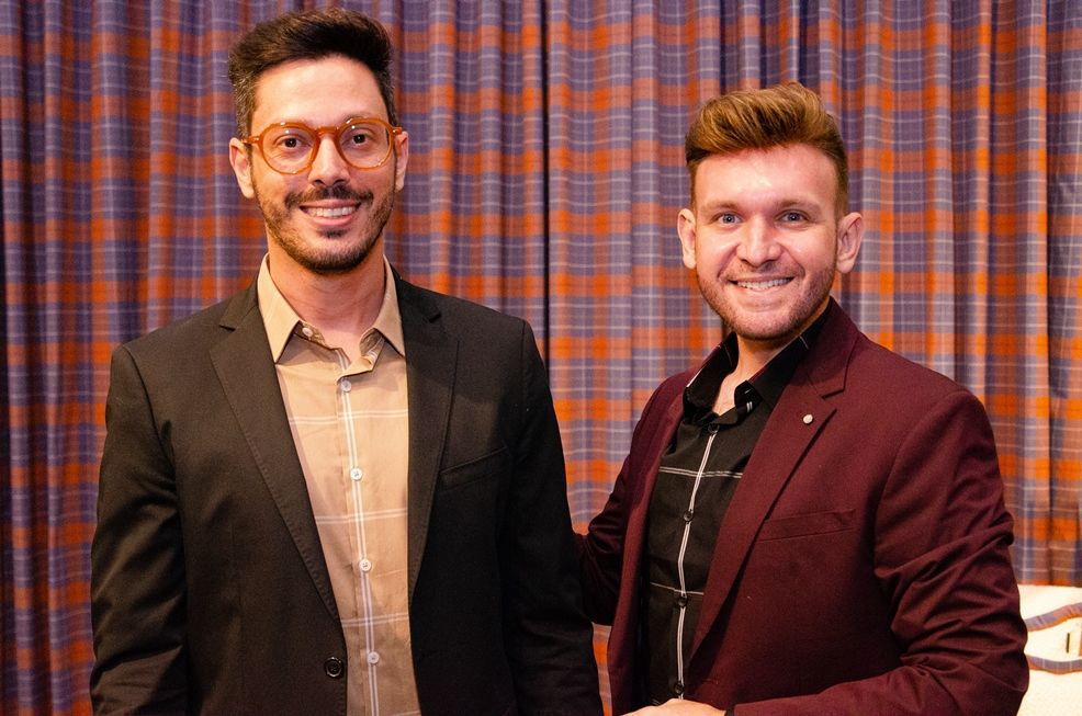 Dois homens de terno apertam as mãos em frente a uma cortina xadrez.