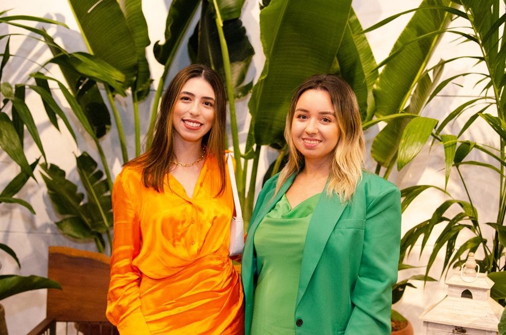 Duas mulheres estão de pé, uma ao lado da outra, em frente a uma planta.