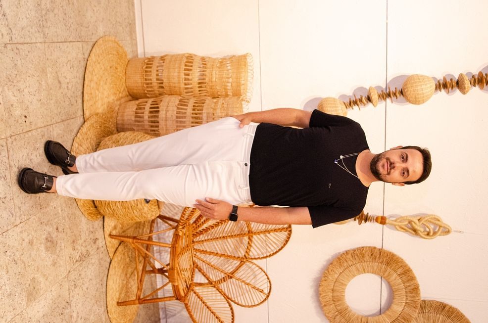 Um homem de camisa preta e calça branca está de pé ao lado de uma cadeira de vime.