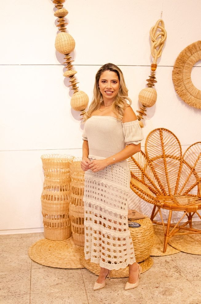 Uma mulher de vestido branco está parada em frente a uma cadeira de vime.