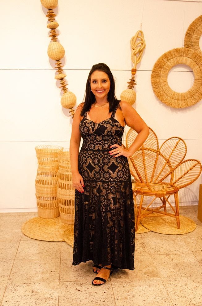 Uma mulher com um vestido de renda preta está parada em frente a cadeiras de vime.