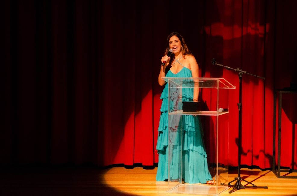 Uma mulher de vestido azul está de pé em um pódio segurando um microfone.