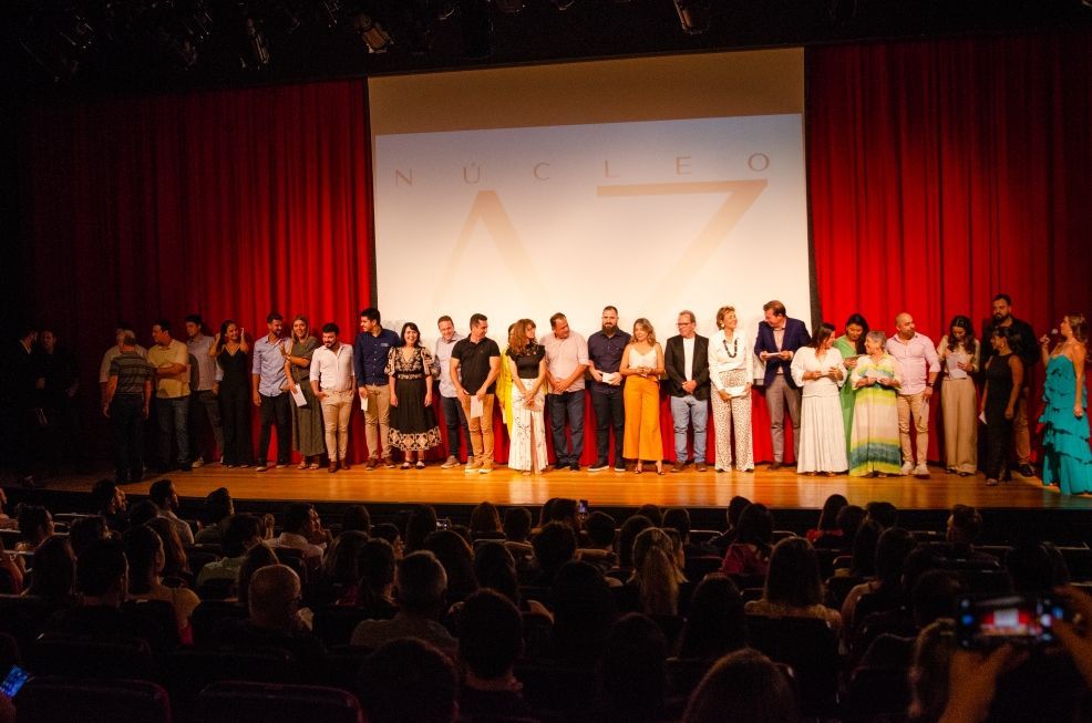 Um grupo de pessoas está em pé em um palco em frente a uma tela grande.