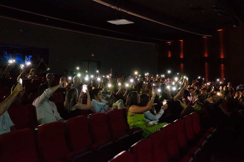 Uma multidão de pessoas está sentada em um teatro segurando seus celulares no ar.