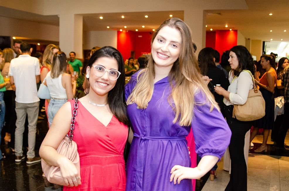 Duas mulheres em vestidos vermelhos e roxos estão posando para uma foto em uma sala lotada.