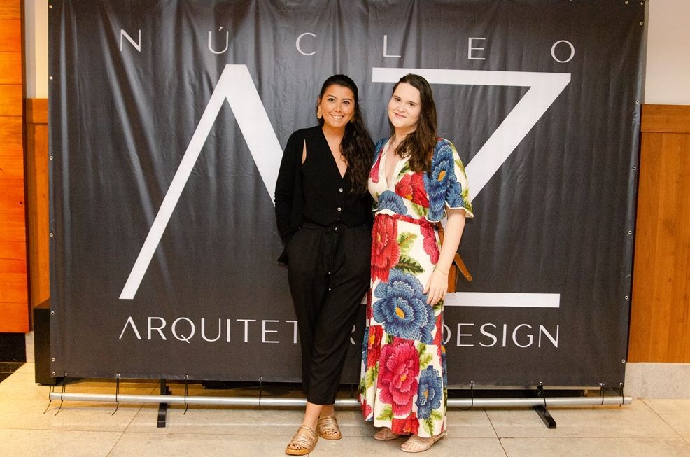 Duas mulheres posam para uma foto em frente a um banner que diz nucleo arquiteto design.