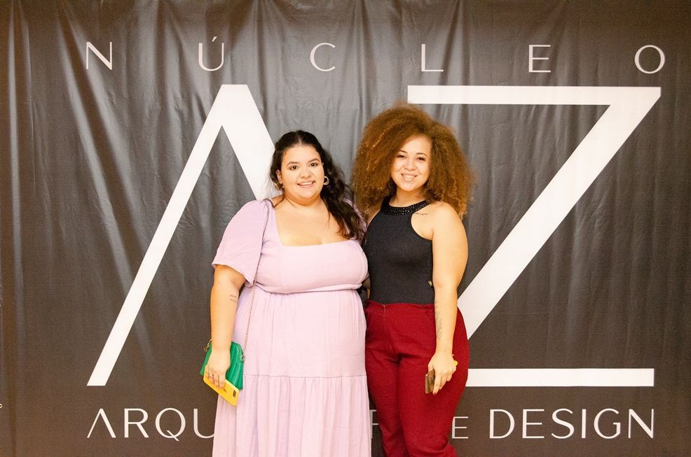 Duas mulheres posam para uma foto em frente a uma placa que diz nucleo arquitetura design.