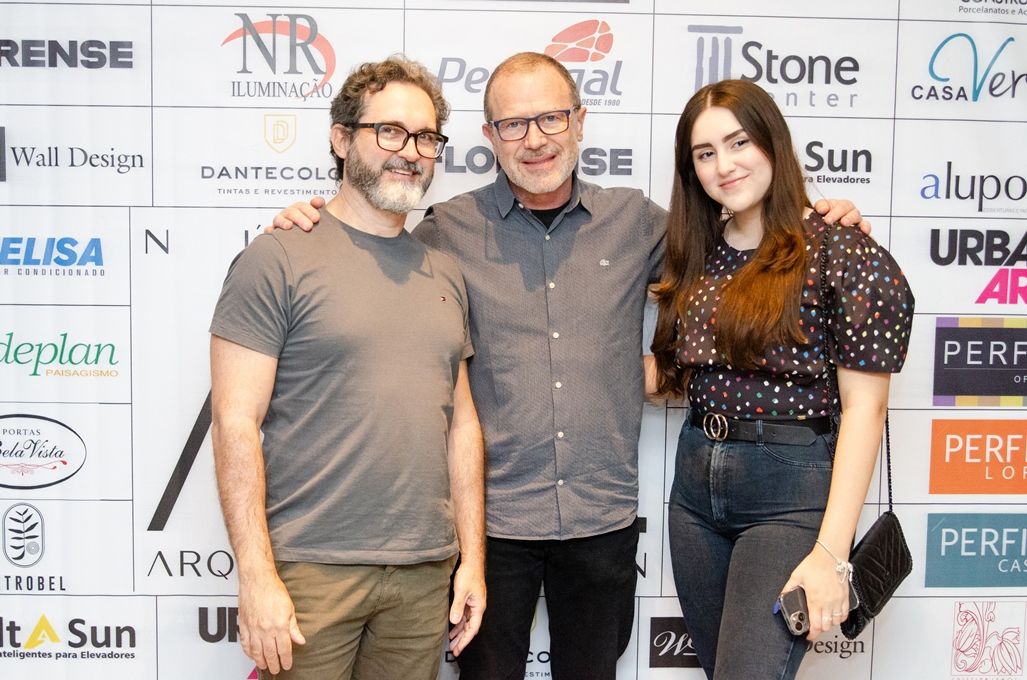 Três pessoas estão posando para uma foto em frente a uma parede com logotipos.