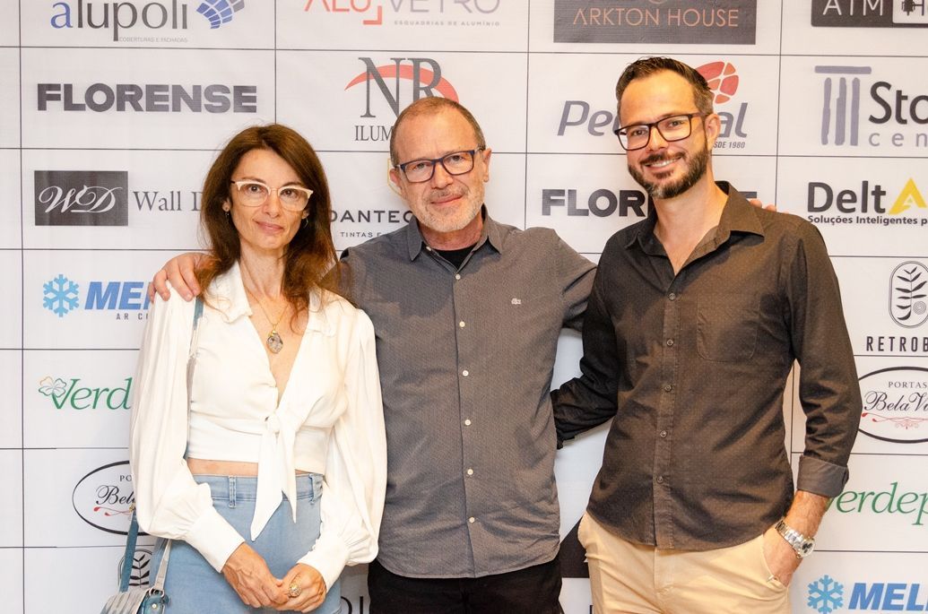 Três pessoas estão posando para uma foto em frente a uma parede com logotipos.