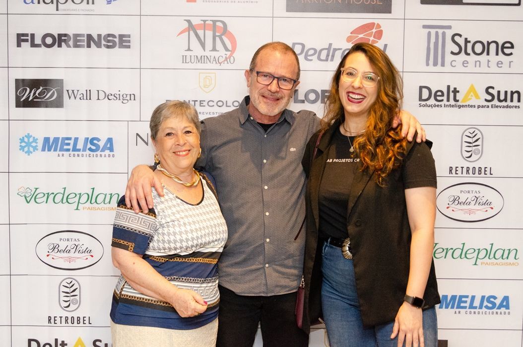 Um homem e duas mulheres estão posando para uma foto em frente a uma parede com logotipos.