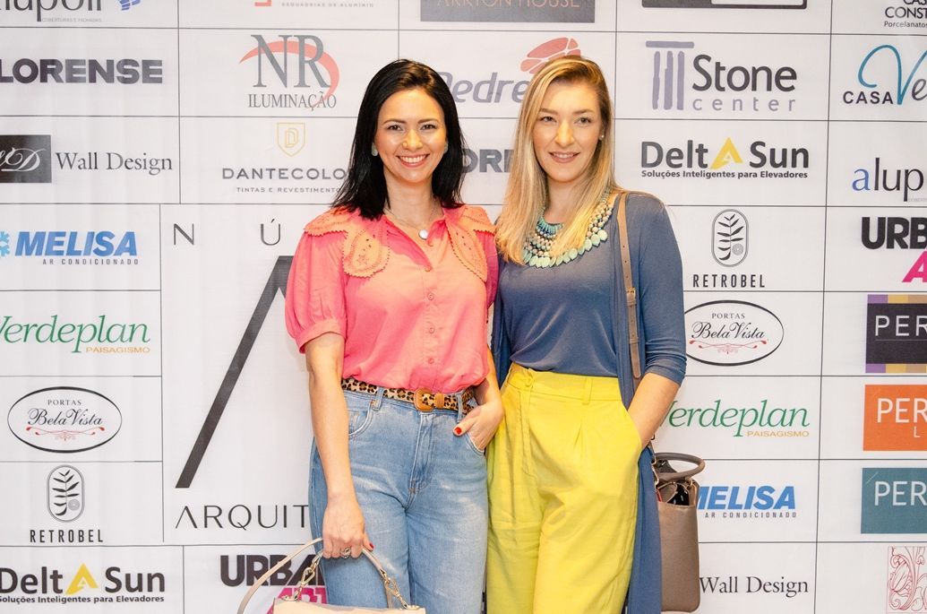 Duas mulheres estão posando para uma foto em frente a uma parede de logotipos.
