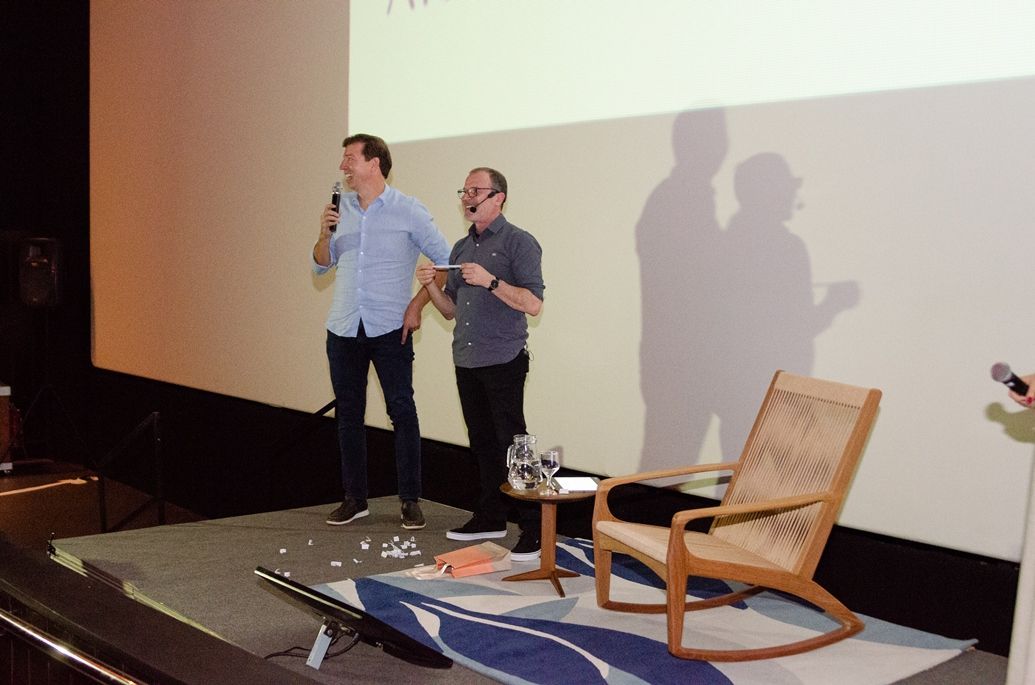 Dois homens estão em pé em um palco falando em microfones enquanto uma cadeira está na frente deles.