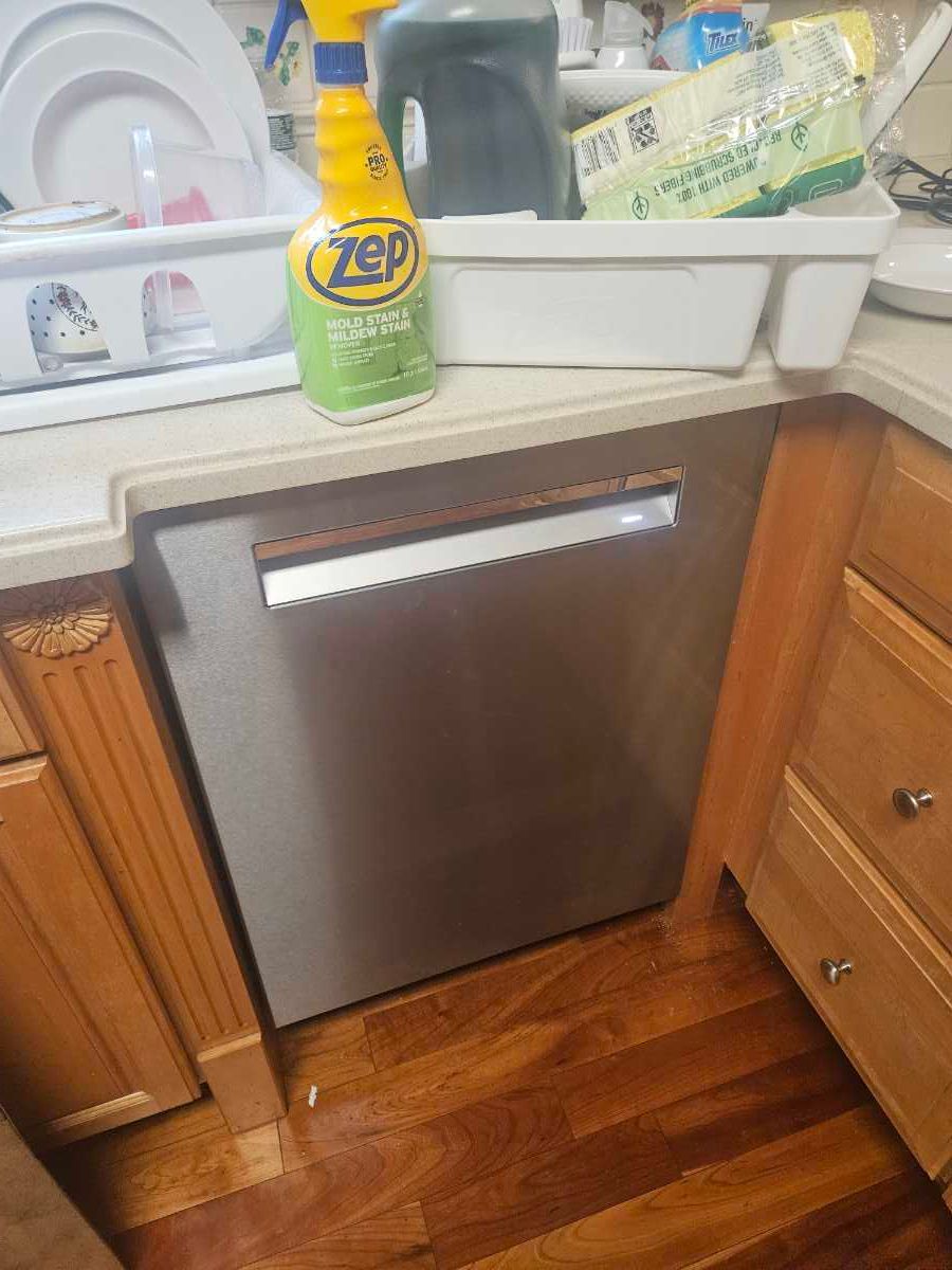 Stainless steel dishwasher with open door, showing racks. Kitchen setting with wood cabinets and white backsplash.