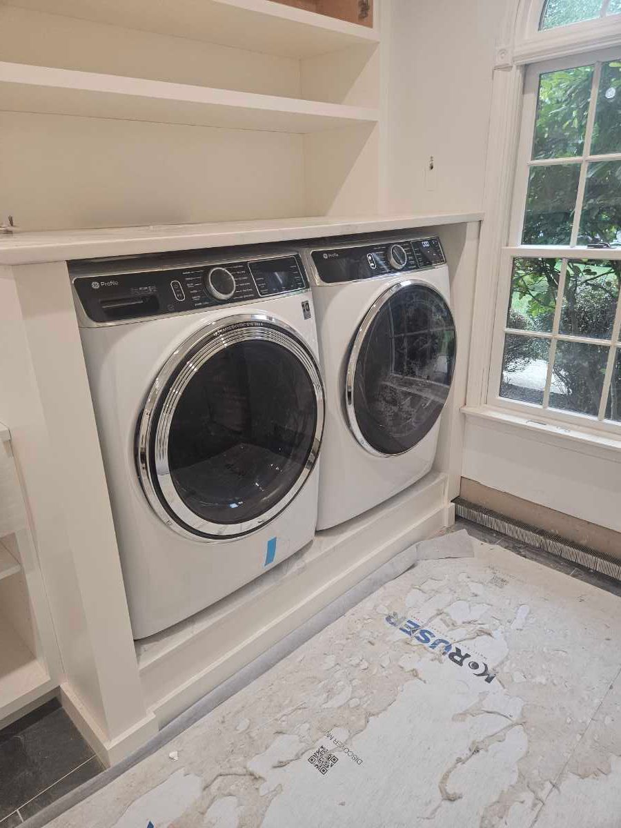 A person in overalls repairs a washing machine in a tiled bathroom, with tools nearby.