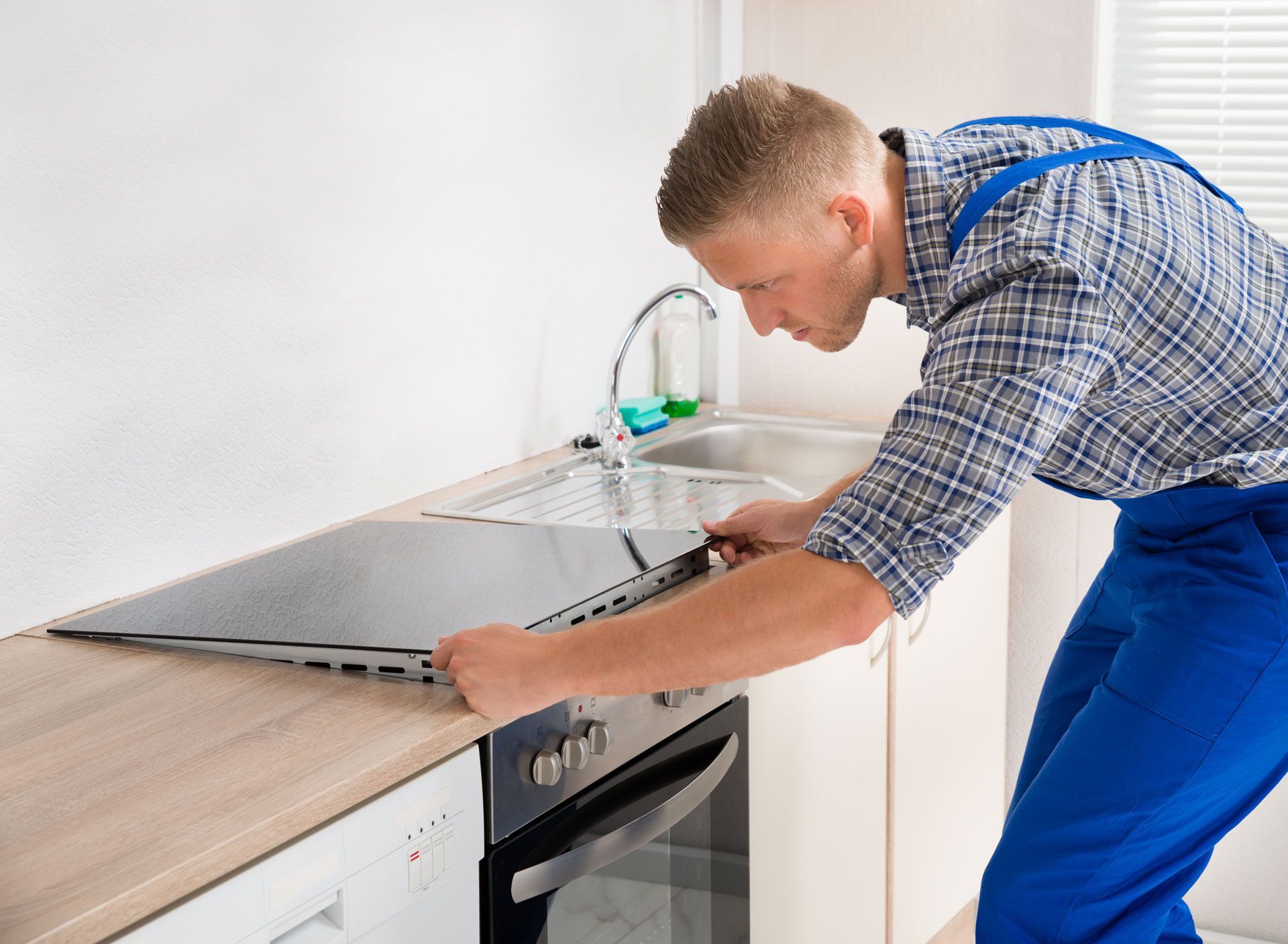 Man in blue overalls installing a black stovetop in a kitchen.