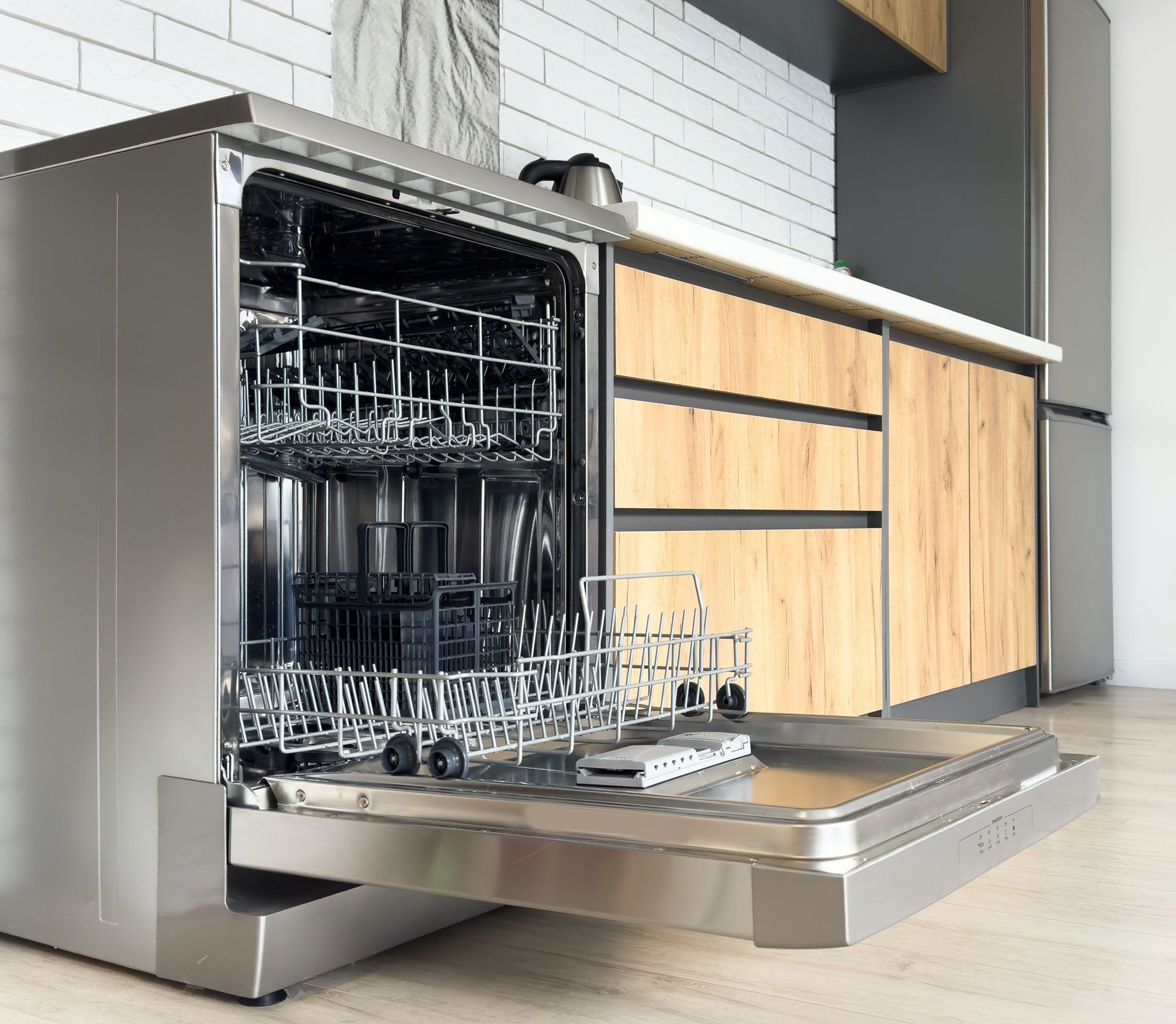 Stainless steel dishwasher with open door, showing racks. Kitchen setting with wood cabinets and white backsplash.