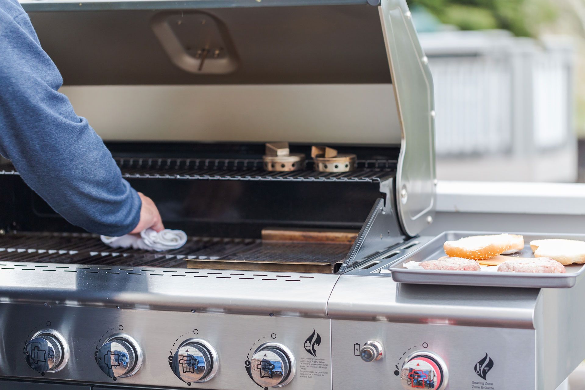 Person grilling on a stainless steel barbecue. Burgers on a tray.