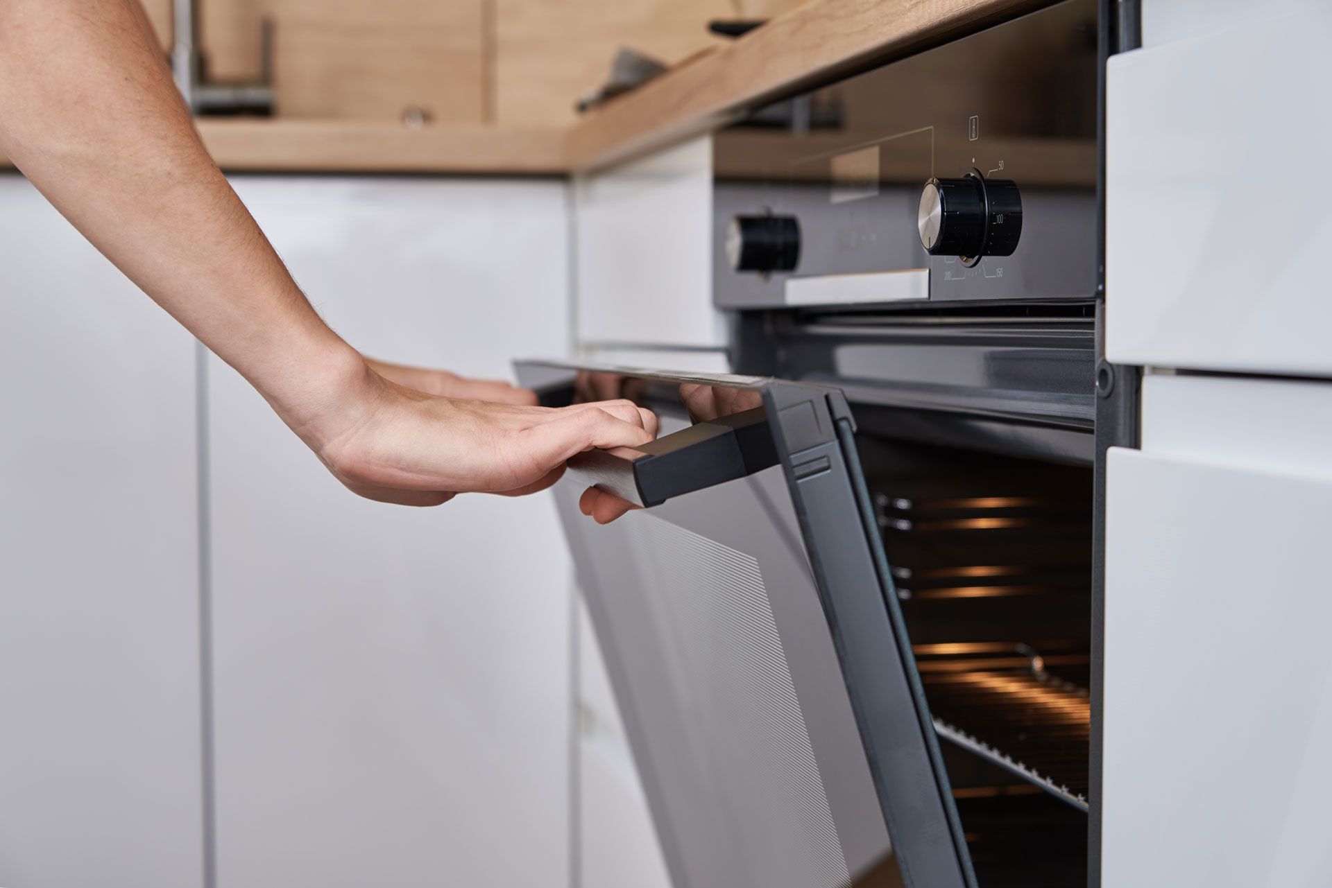 Hand opening a modern oven door in a white kitchen; oven interior glows with light.