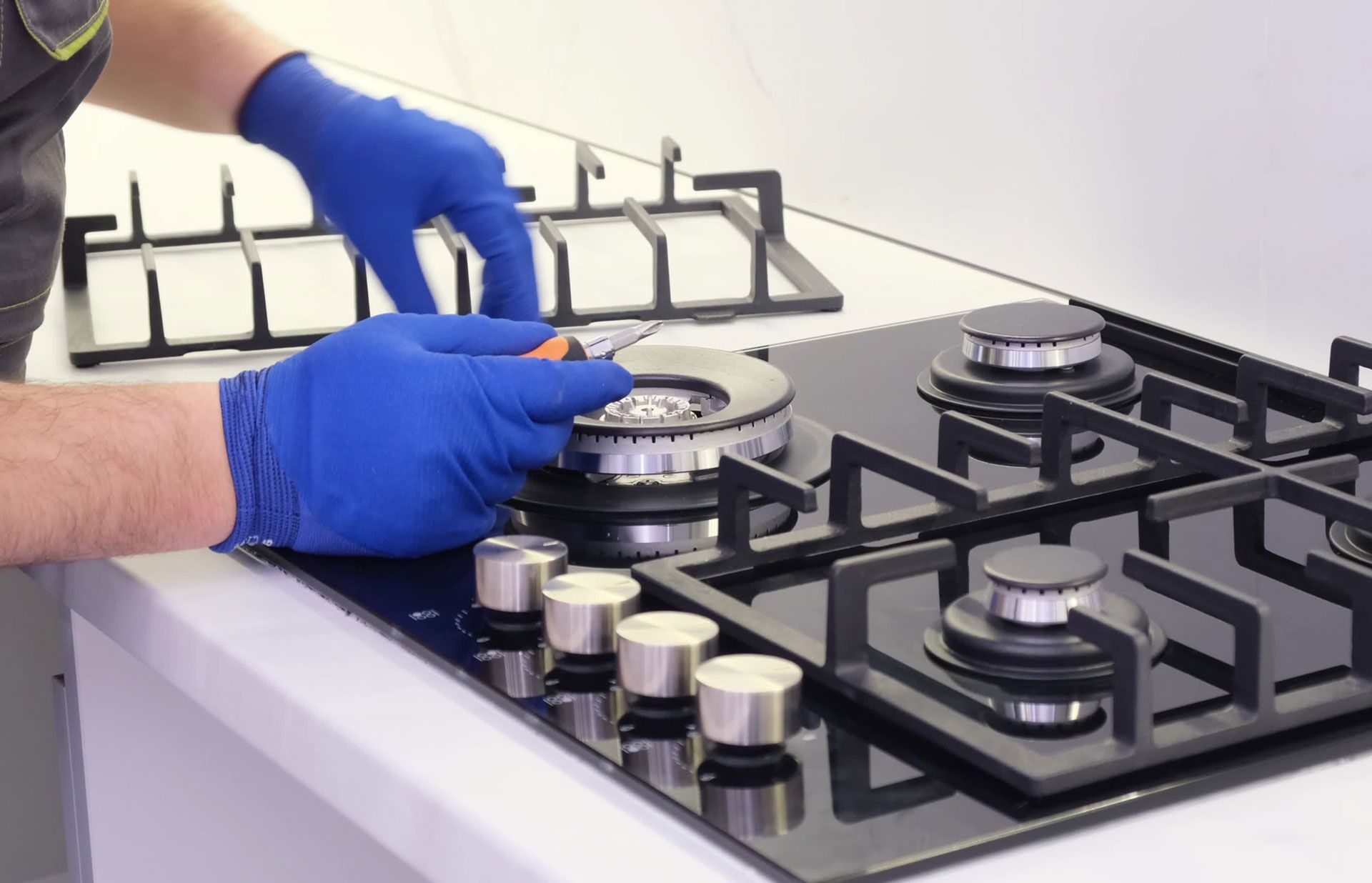 Person in blue gloves fixing a black gas stovetop with a tool.