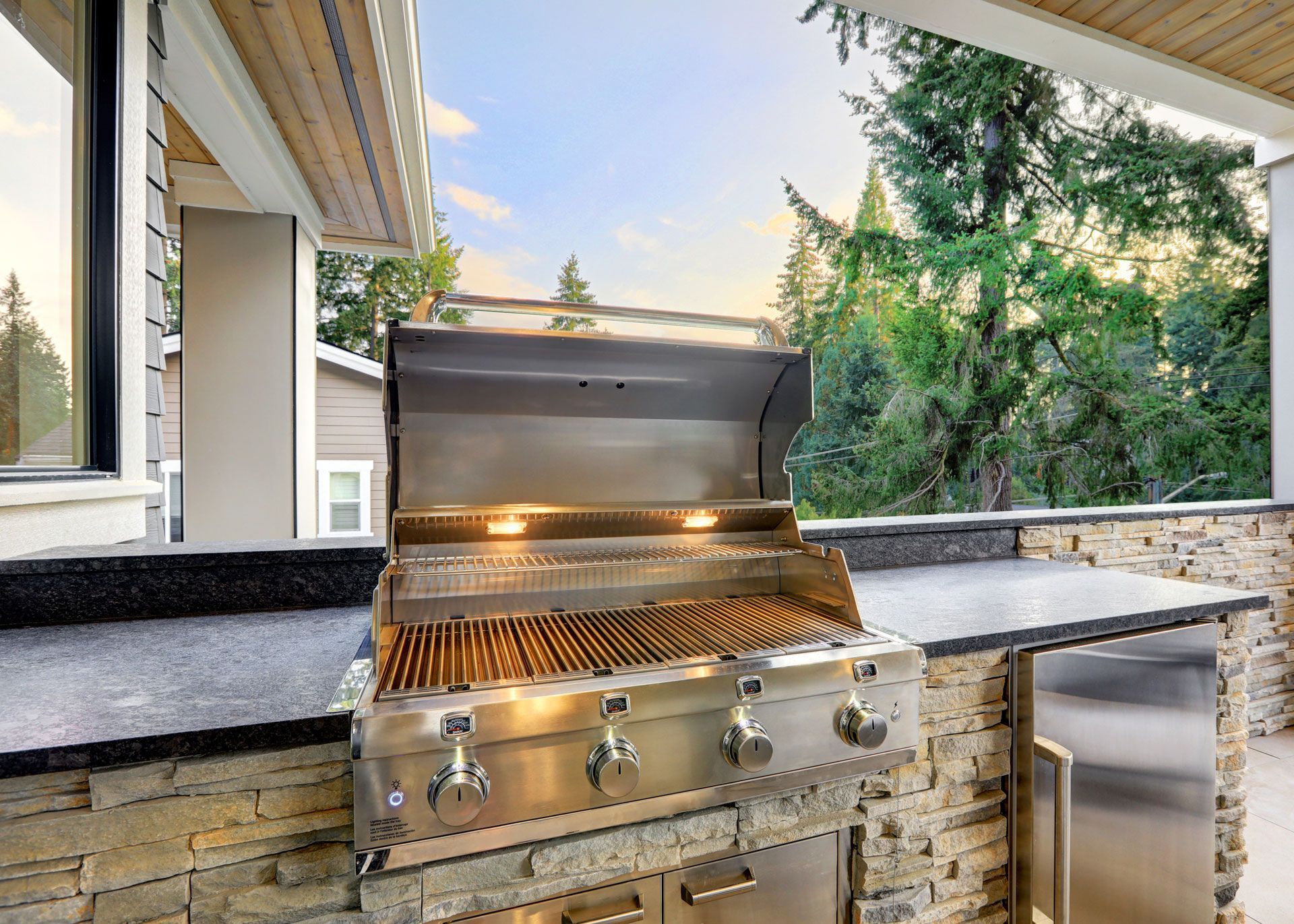 Stainless steel outdoor grill on stone patio with a wooded background.