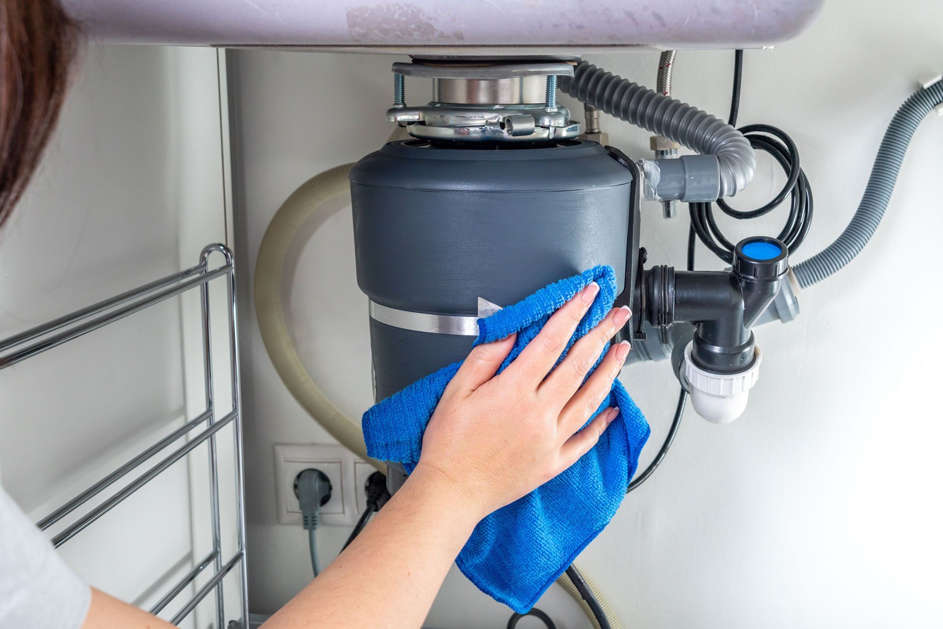 Person wiping a gray garbage disposal under a sink with a blue cloth.