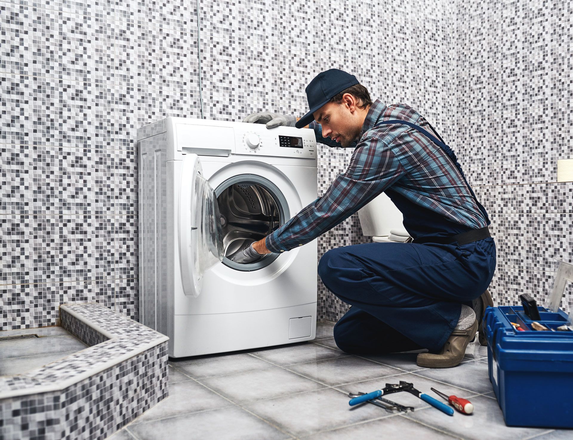 A person in overalls repairs a washing machine in a tiled bathroom, with tools nearby.