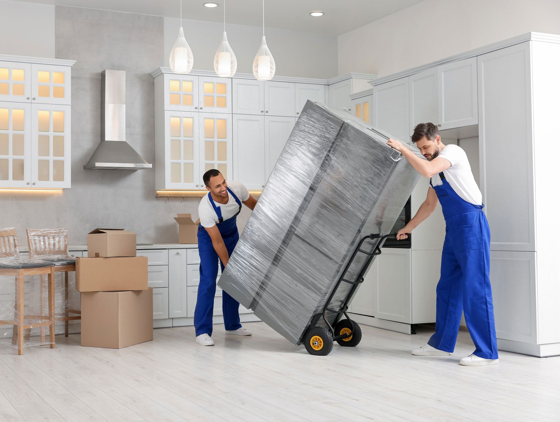Two movers in blue overalls using a hand truck to move a large, wrapped object in a white kitchen.