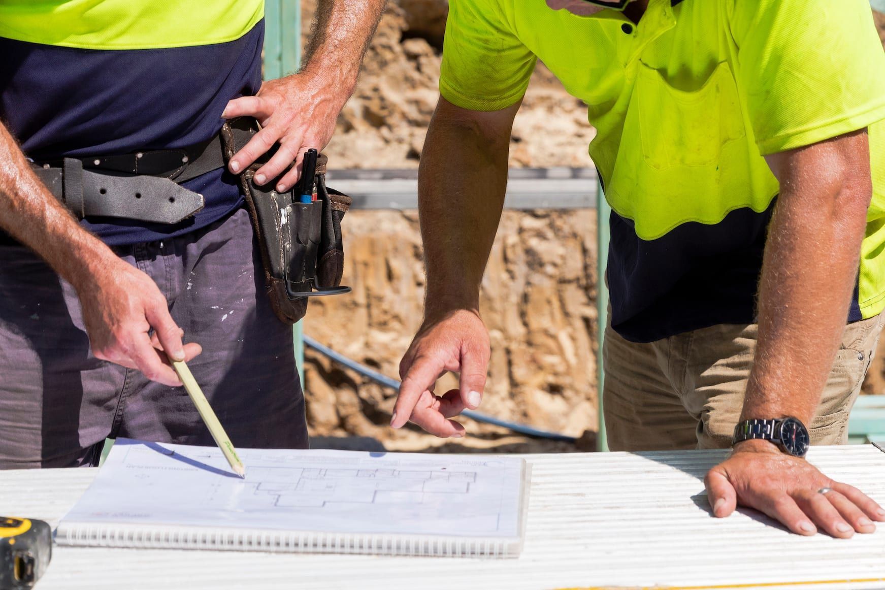 Two construction workers examining blueprints on a table, outdoors. One points with a pen, the other with a finger.