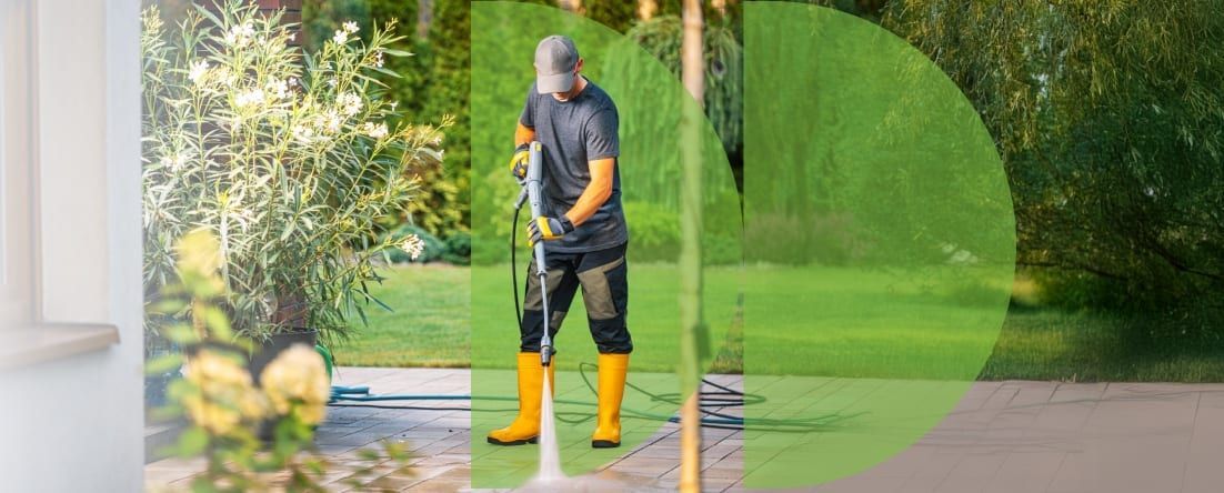 A person in yellow boots power washes a patio, standing on the patio in a yard.