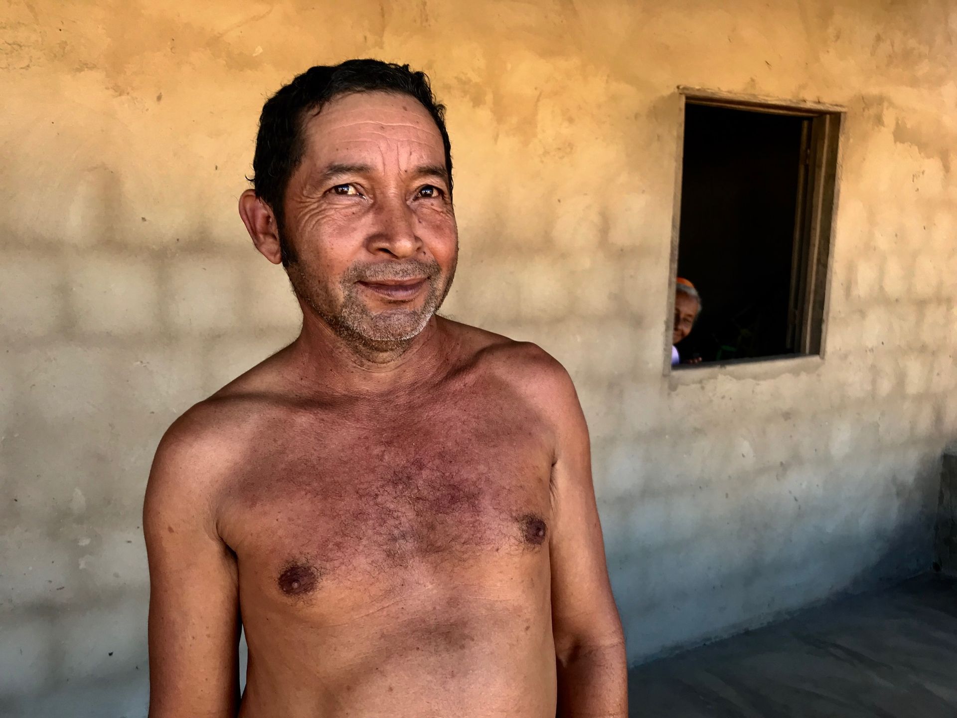 Um homem sem camisa está parado em frente a uma janela e sorrindo.