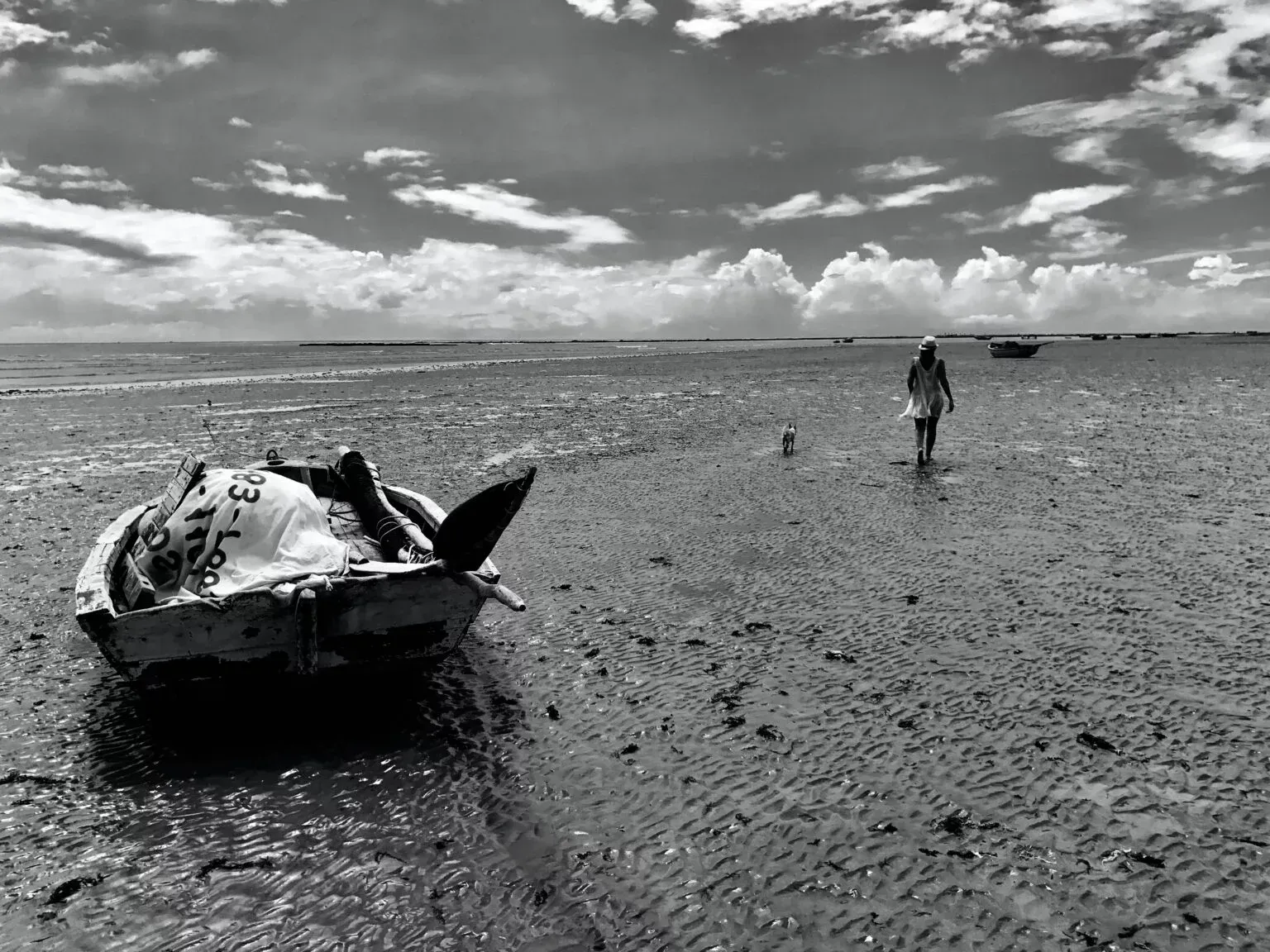 Uma foto em preto e branco de um homem caminhando em uma praia ao lado de um barco.