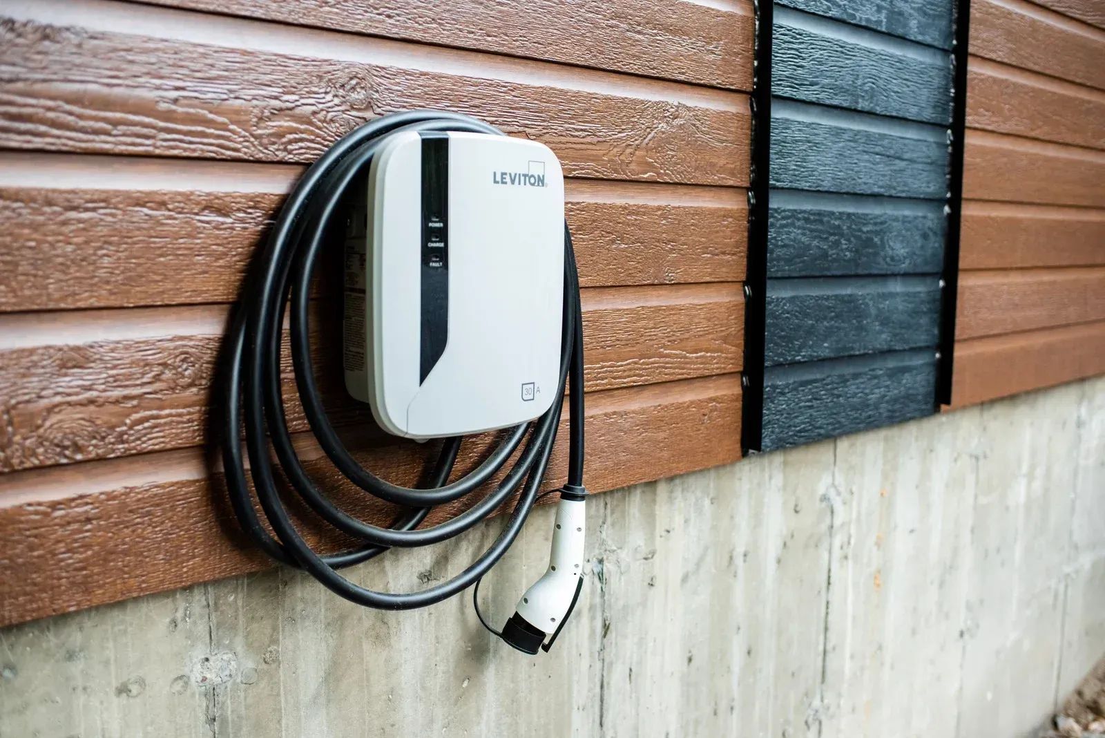 Wall-mounted white EV charger with coiled black cable on brown and black siding above concrete.