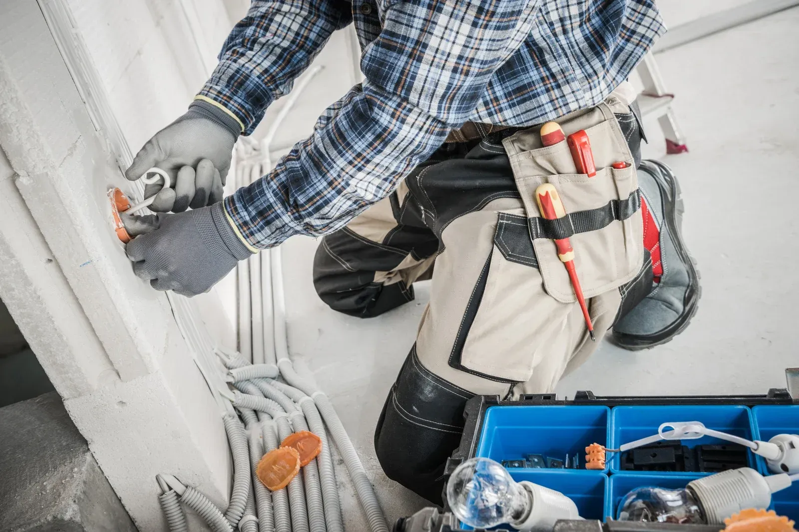 Electrician installing electrical wiring on a wall, wearing gloves and carrying tools in a belt.