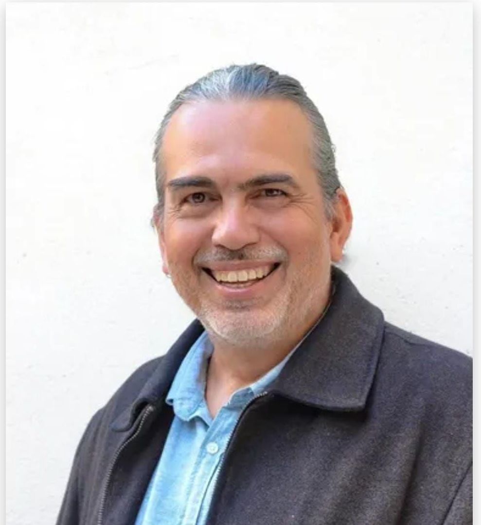 Um homem de cabelos grisalhos  vestindo uma jaqueta e uma camisa azul está sorrindo.