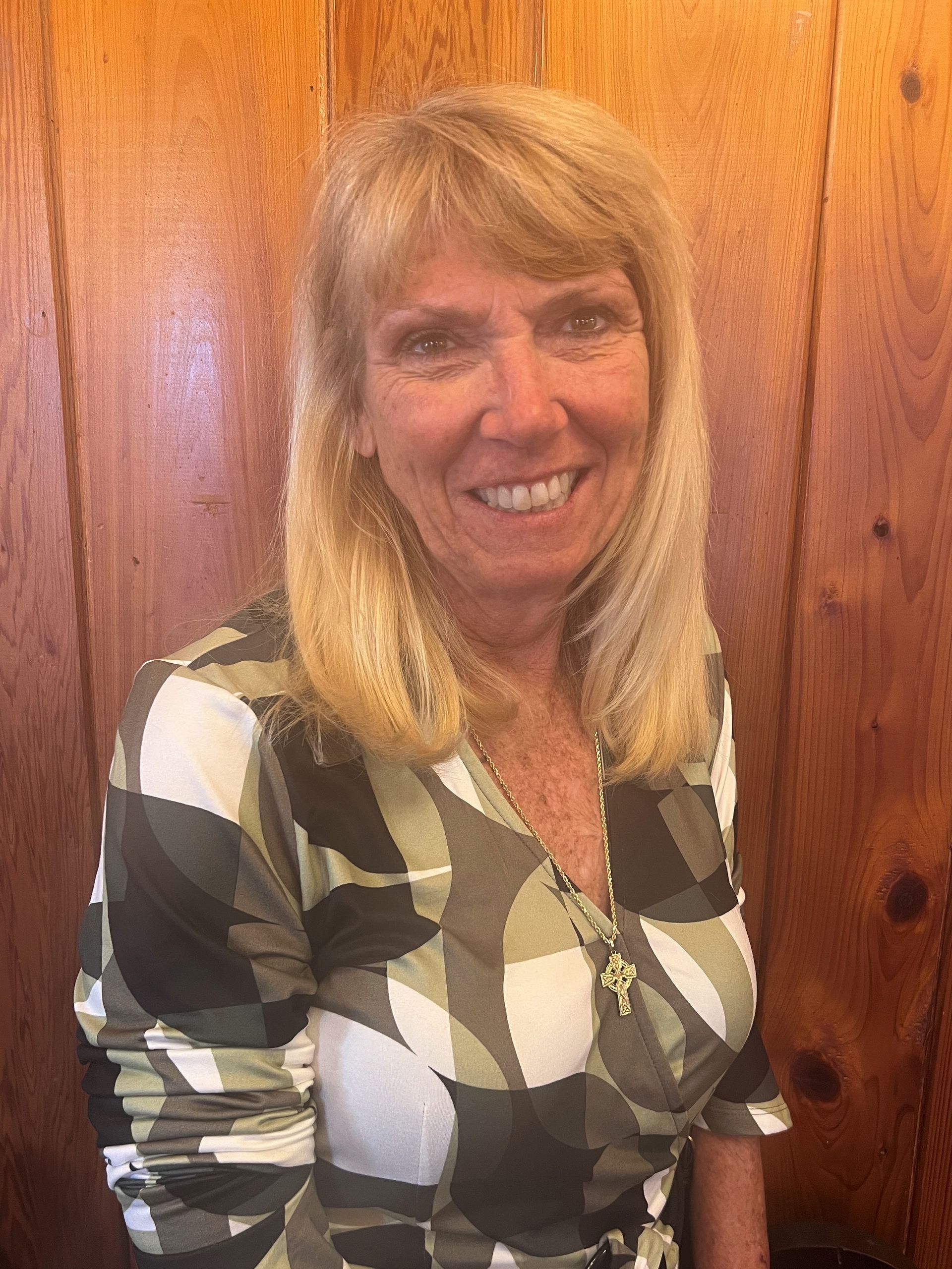 A woman in a camouflage shirt is smiling in front of a wooden wall.
