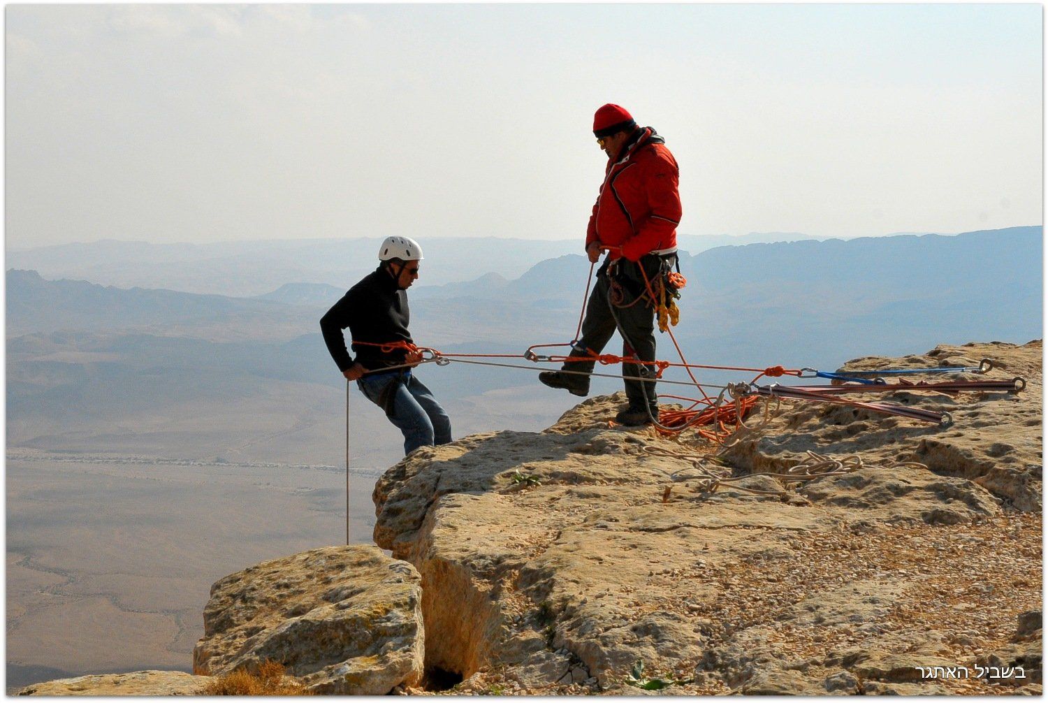 שני גברים עומדים על ראש צוק סלעי עם הרים ברקע