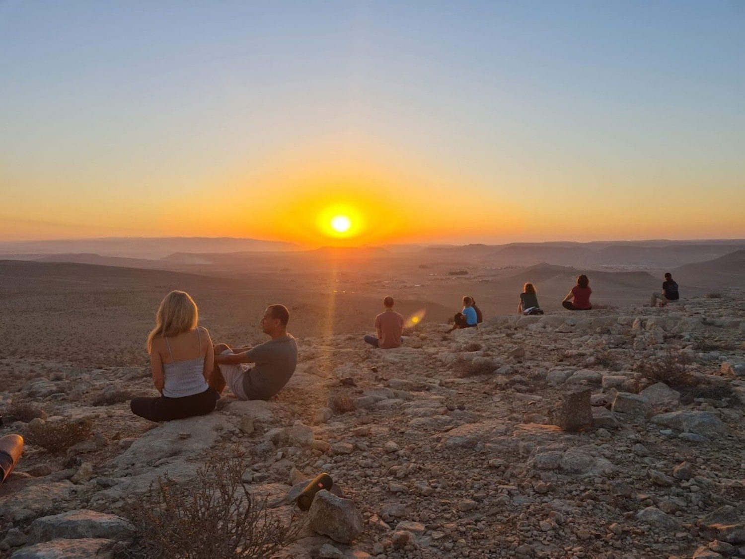קבוצה של אנשים יושבת על ראש גבעה סלעית וצופה בשמש שוקעת.