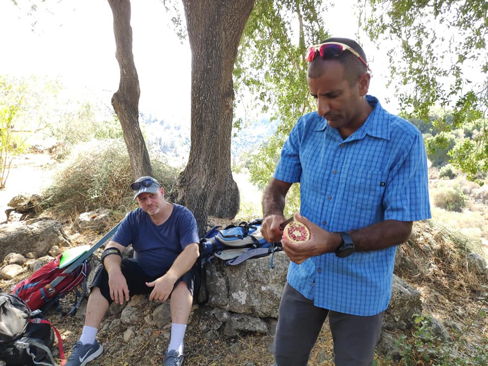 גבר בחולצה כחולה עומד ליד גבר אחר יושב מתחת לעץ.