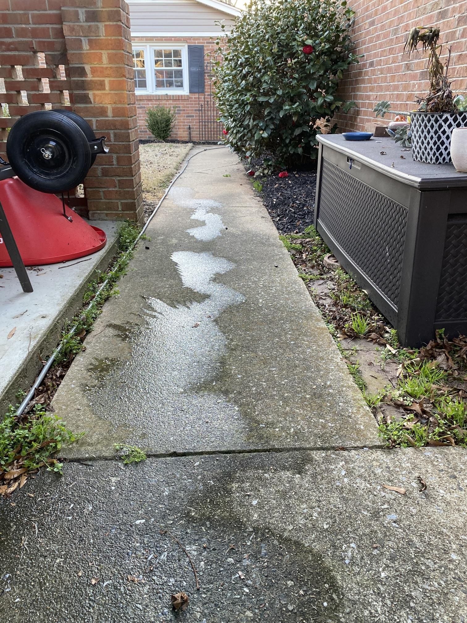 A concrete walkway leading to a brick house with a wheelbarrow in the background.
