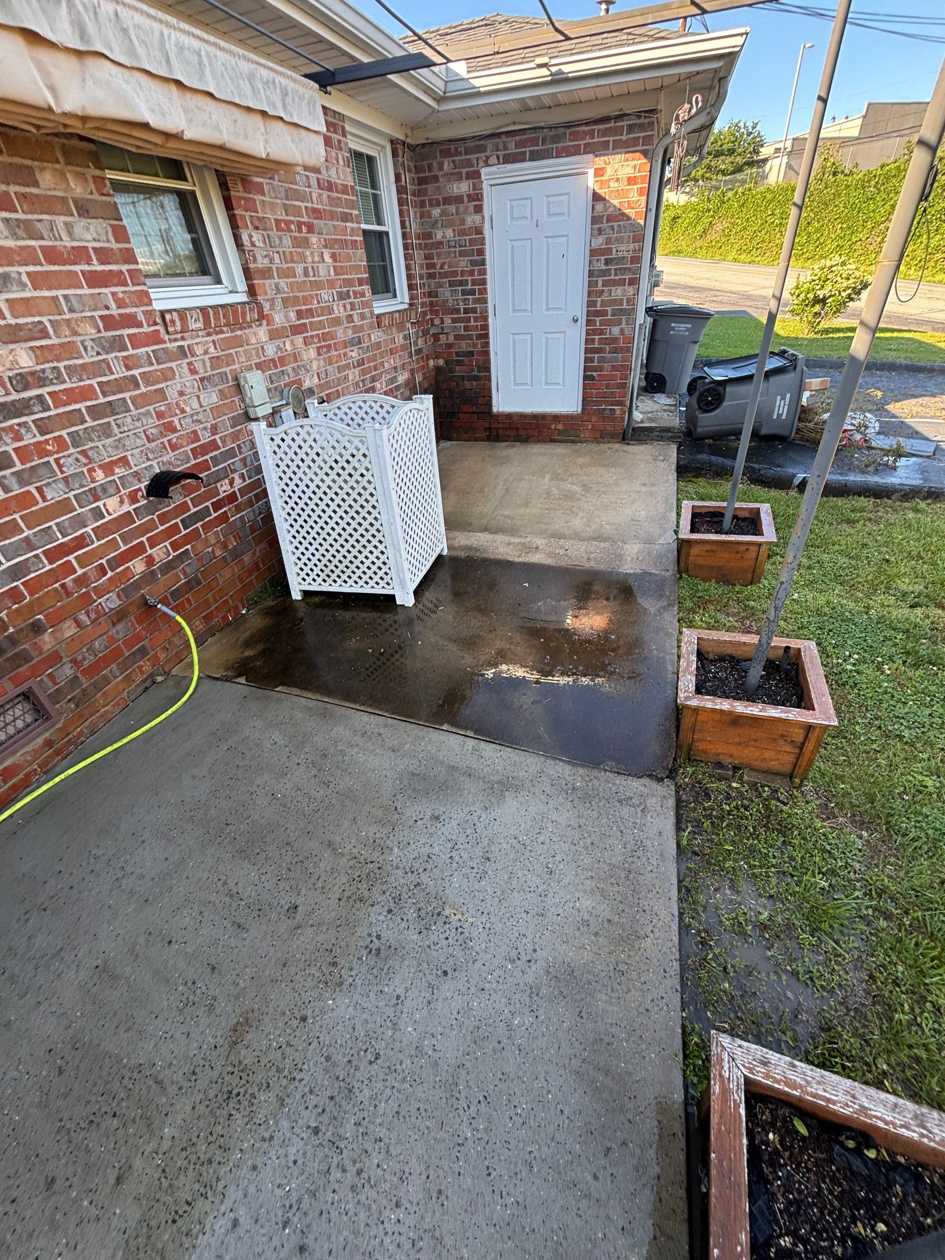 Brick building exterior with wet concrete patio. White lattice bin, brown planters, and a white door are visible.