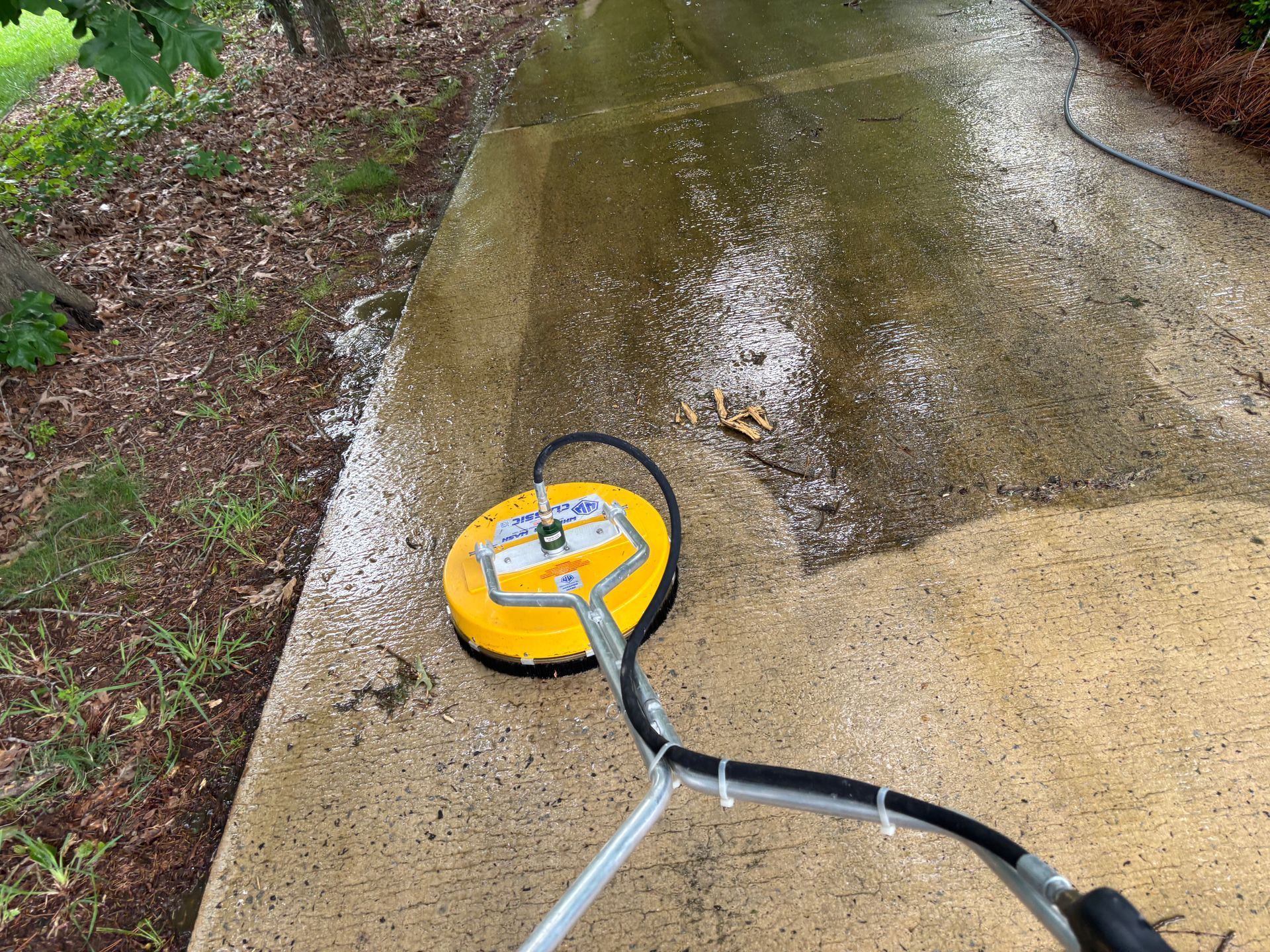 A person using a yellow surface cleaner on a concrete walkway. Wet concrete with white suds is visible.