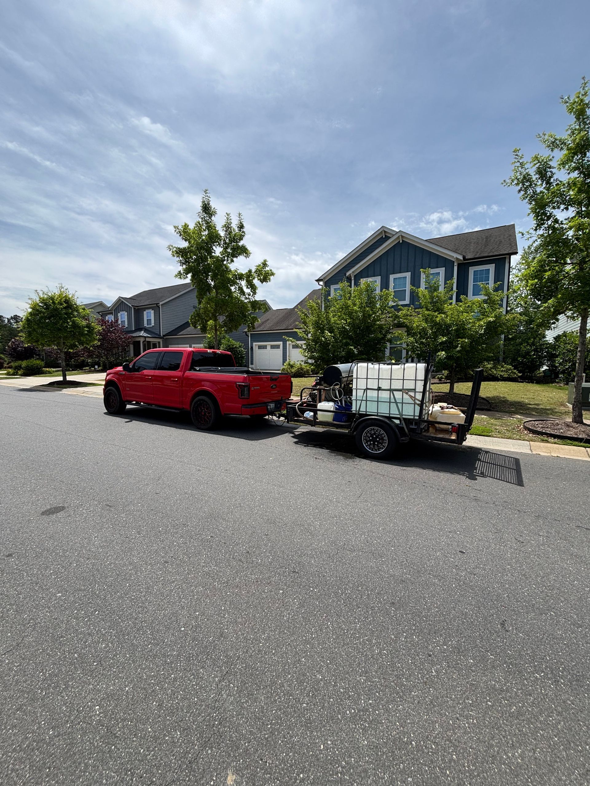 Red truck towing a water tank trailer on a paved street in front of a blue house.