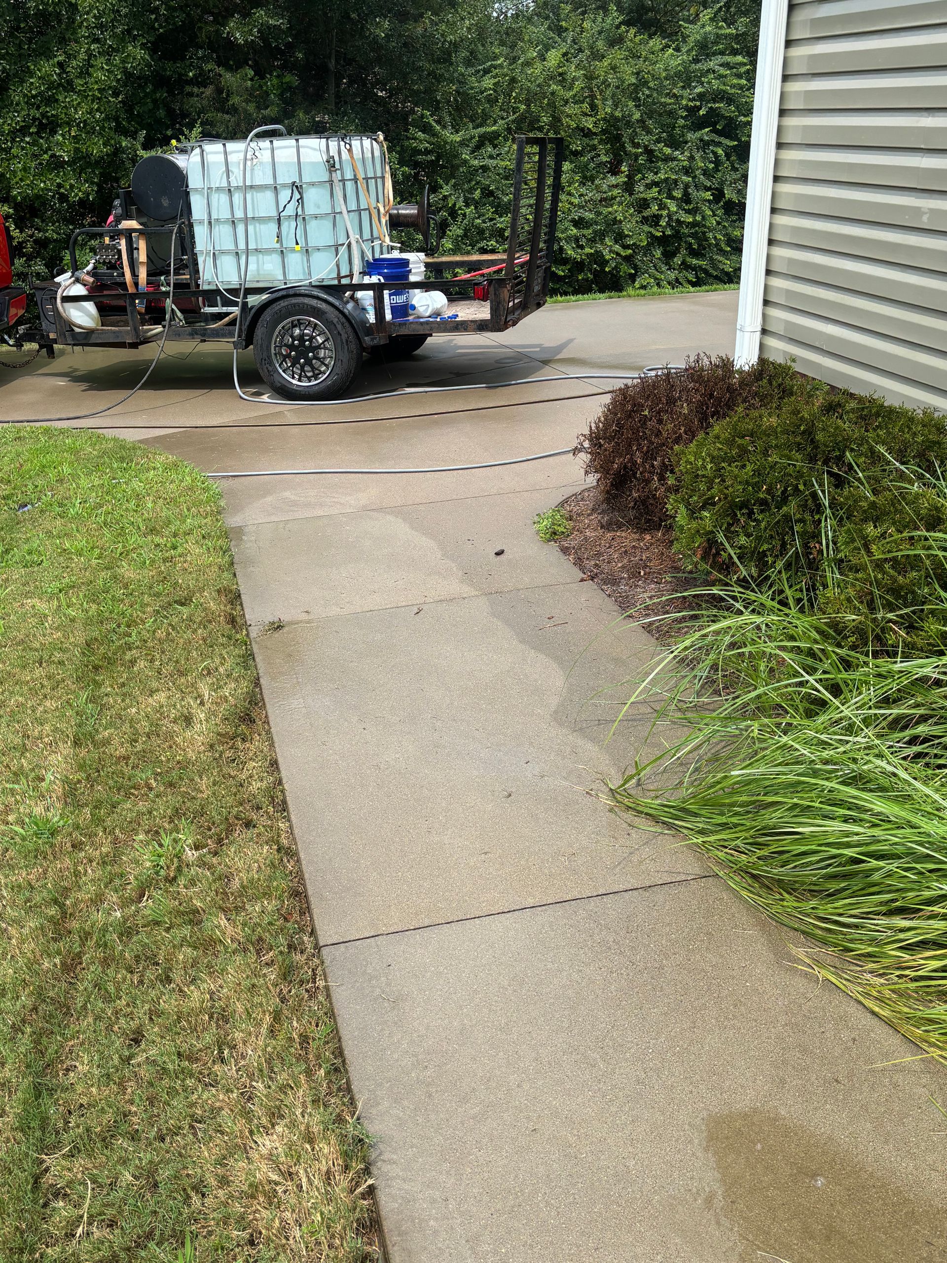 A trailer with a large water tank spraying a concrete walkway next to grass and a building.